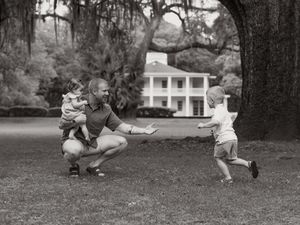 Black and white photo of a father crouching on grass, holding a toddler while another child runs toward him near a white house.