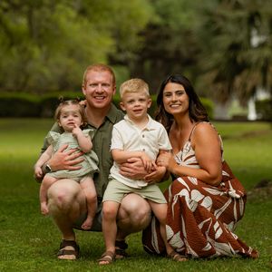Family of four smiling outdoors on green grass, parents with two young children posing for a portrait in a lush park setting.