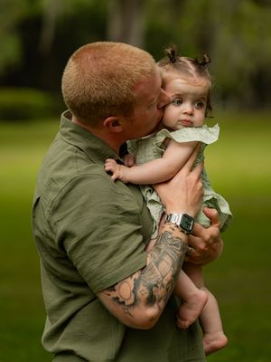 Tattooed father with short red hair kisses baby girl in green dress outdoors.