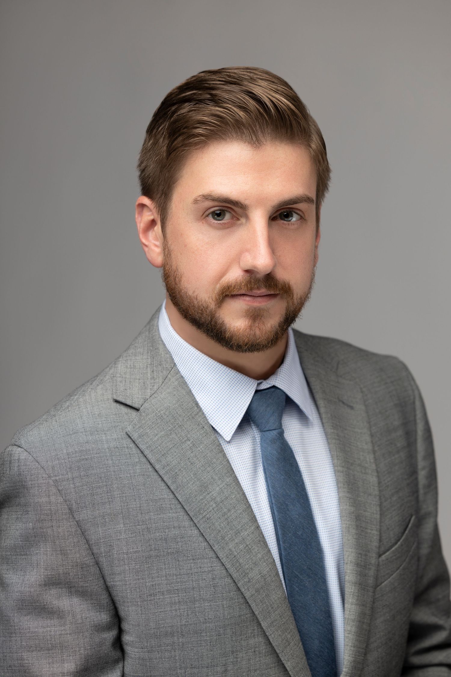 Business headshot featuring person in gray suit and blue tie against neutral background.