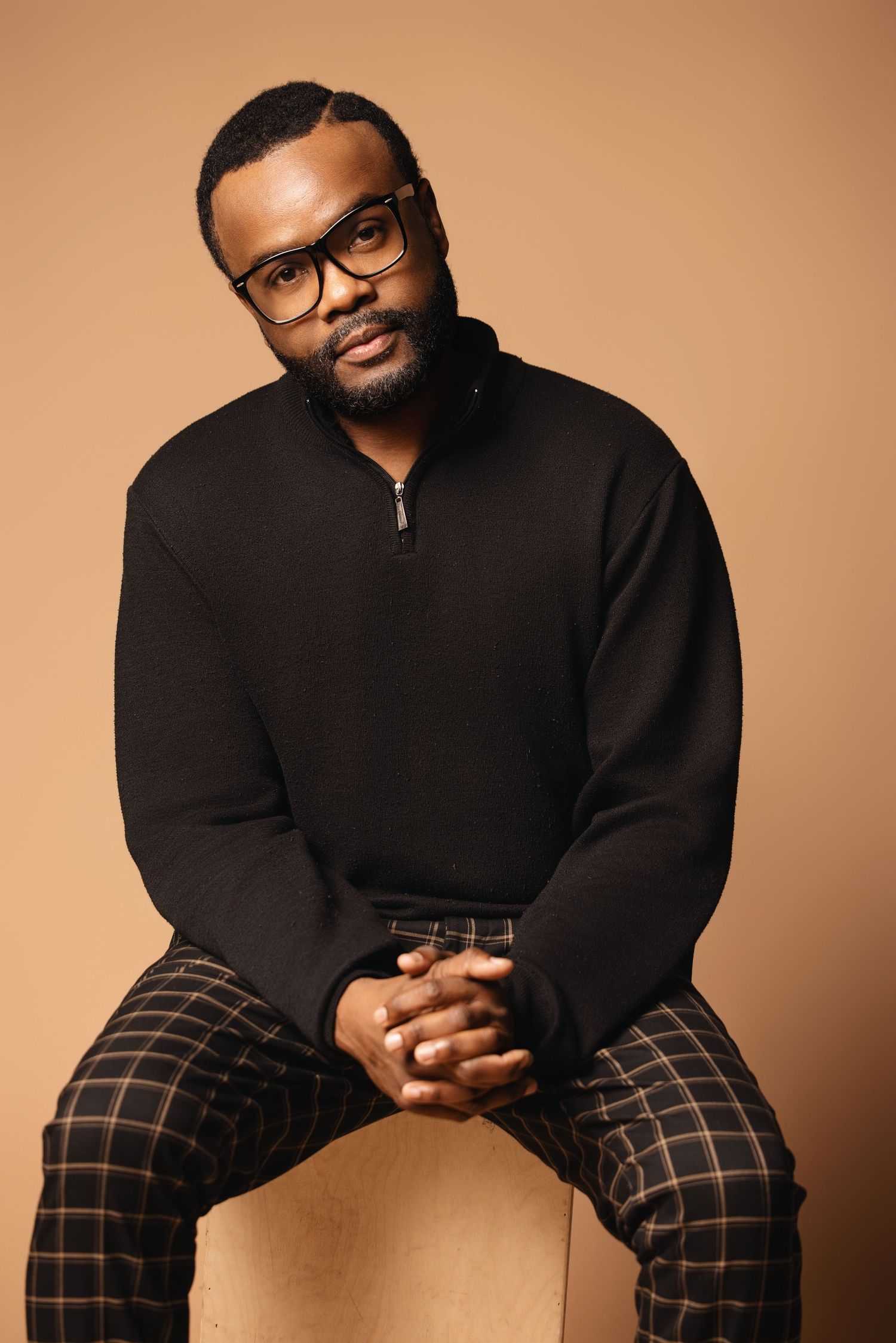 Person wearing black sweater and plaid pants seated casually against warm brown background.