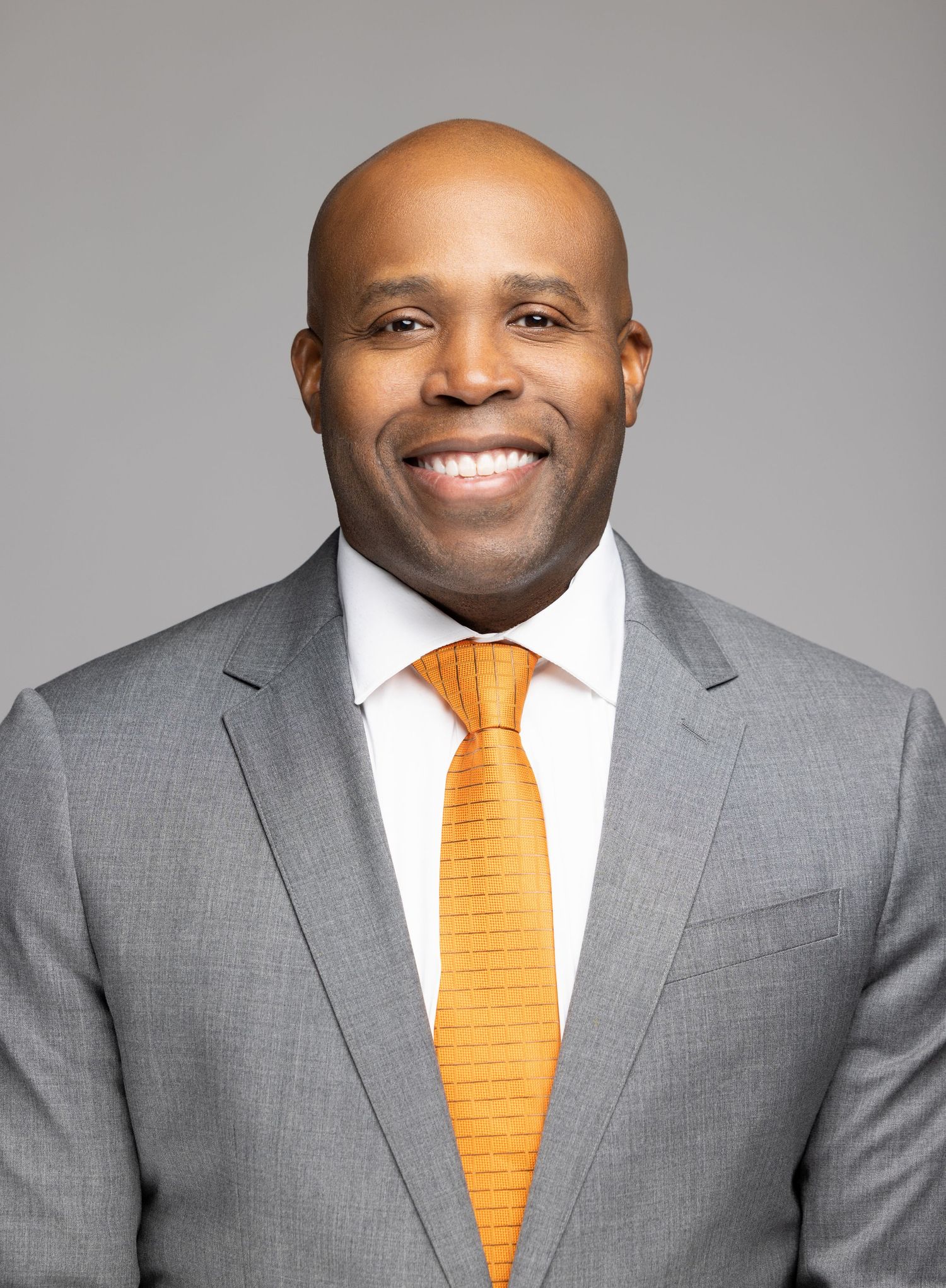 Professional headshot of someone in a grey suit and bright orange tie smiling warmly against a grey background.