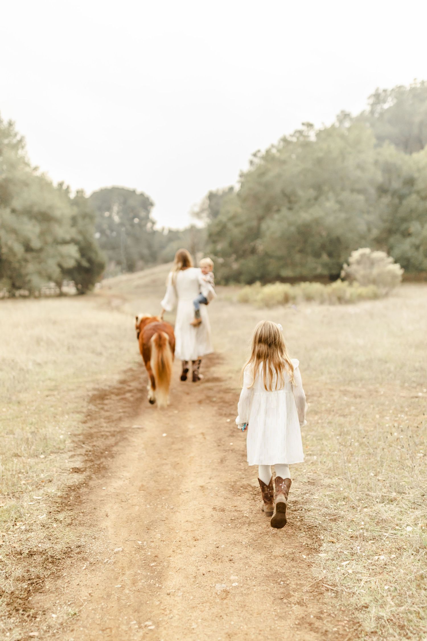 A young girl kisses her smiling mother on the cheek outdoors in a golden field, both wearing white outfits.