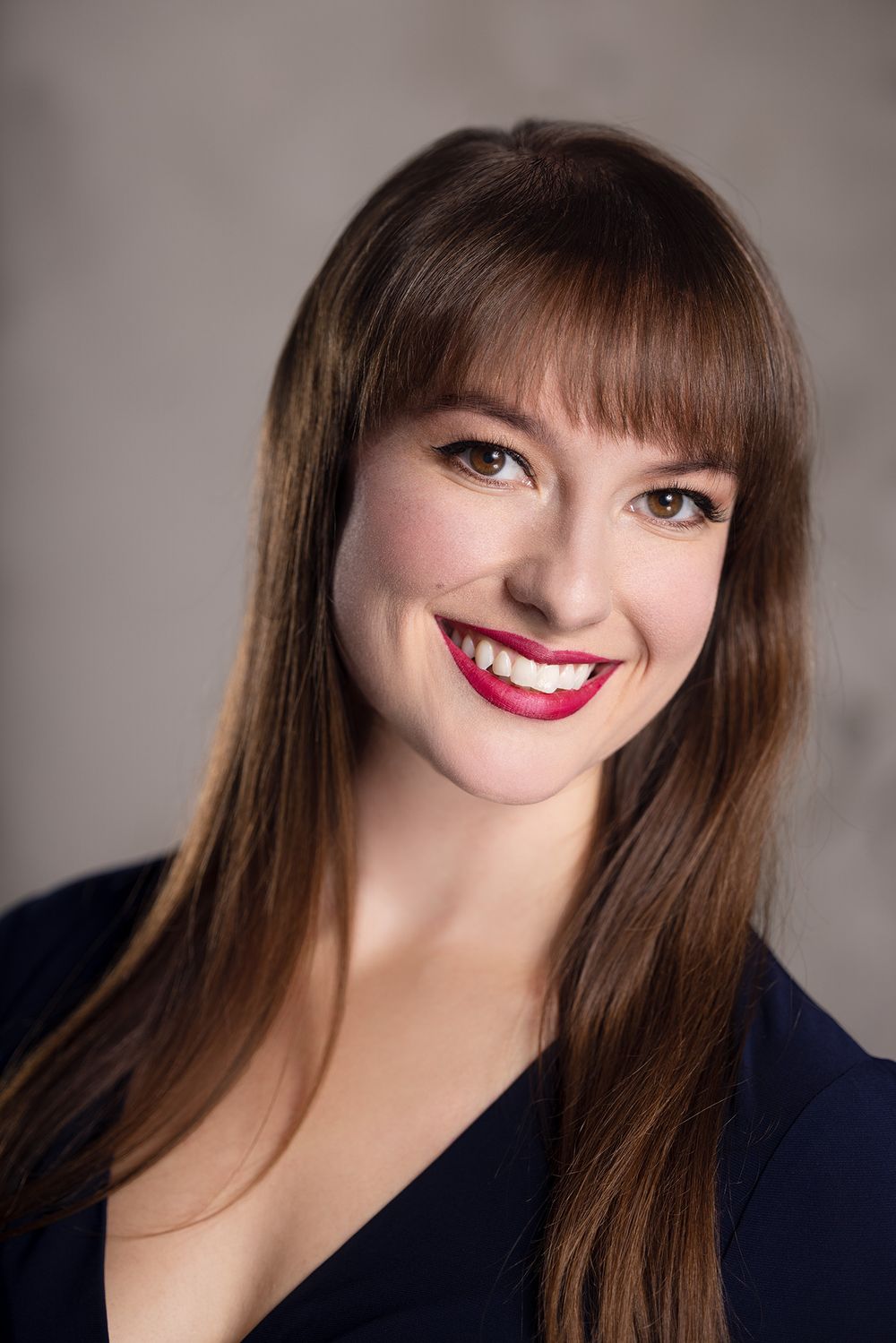 Smiling young woman with long brown hair and red lips wearing a navy top against a gray background. corporate headshot by Gerardo & Co.
