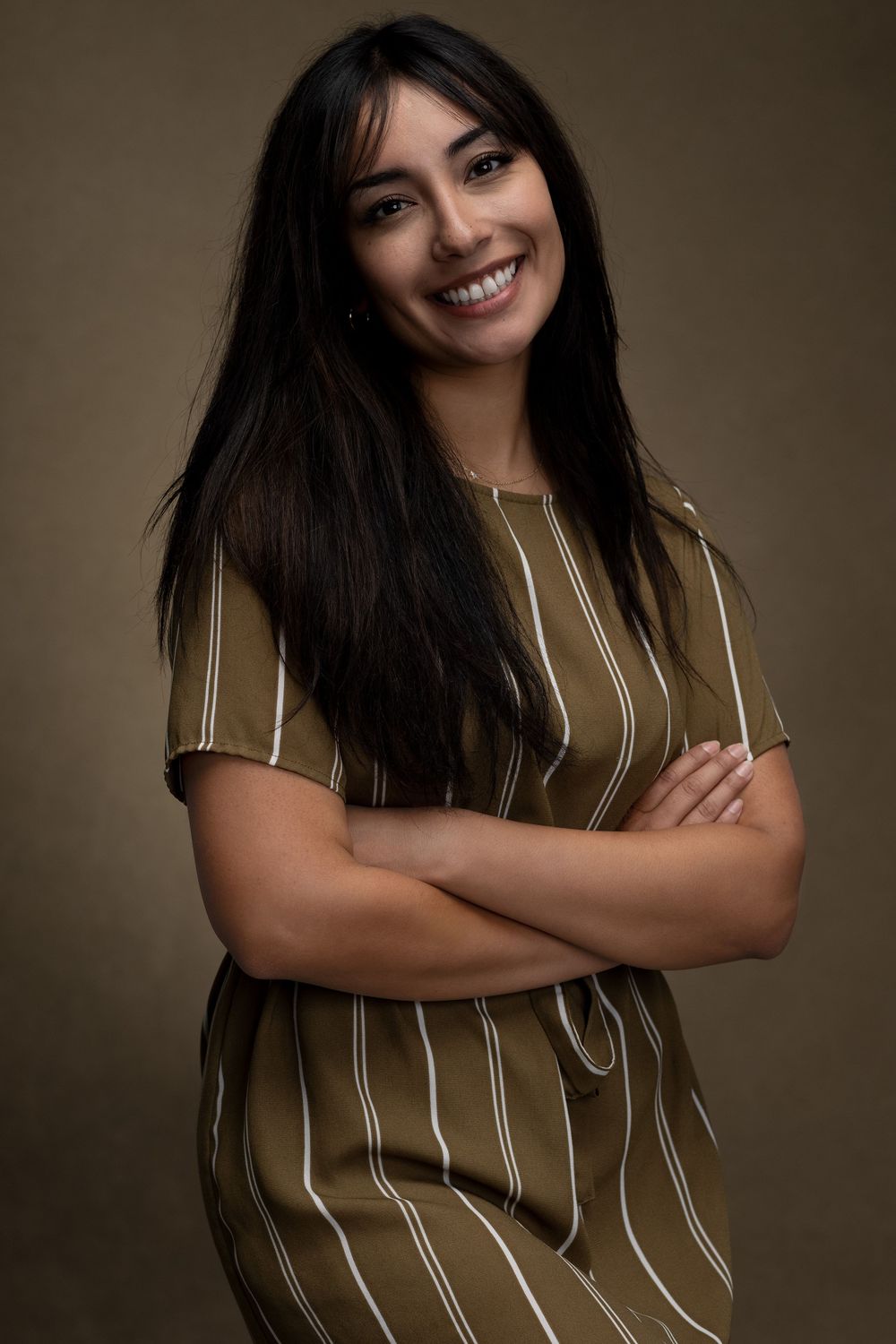 Smiling young woman with long dark hair, arms crossed, wearing an olive striped dress against a dark background. Minneapolis photographer
