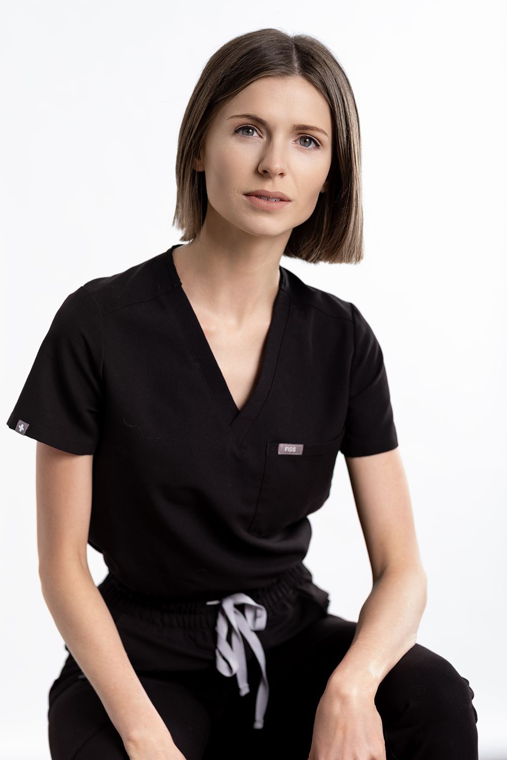 Woman in black medical scrubs uniform sitting against white background, wearing v-neck top and drawstring pants. Gerardoco.com