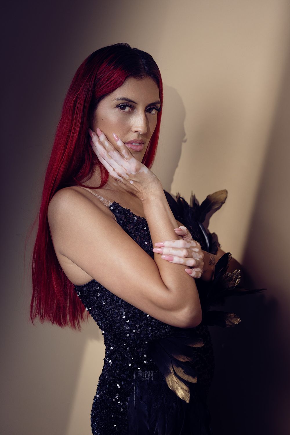 A woman with long red hair in a black sequined dress poses elegantly holding dark feathers against a neutral background. studio photographer