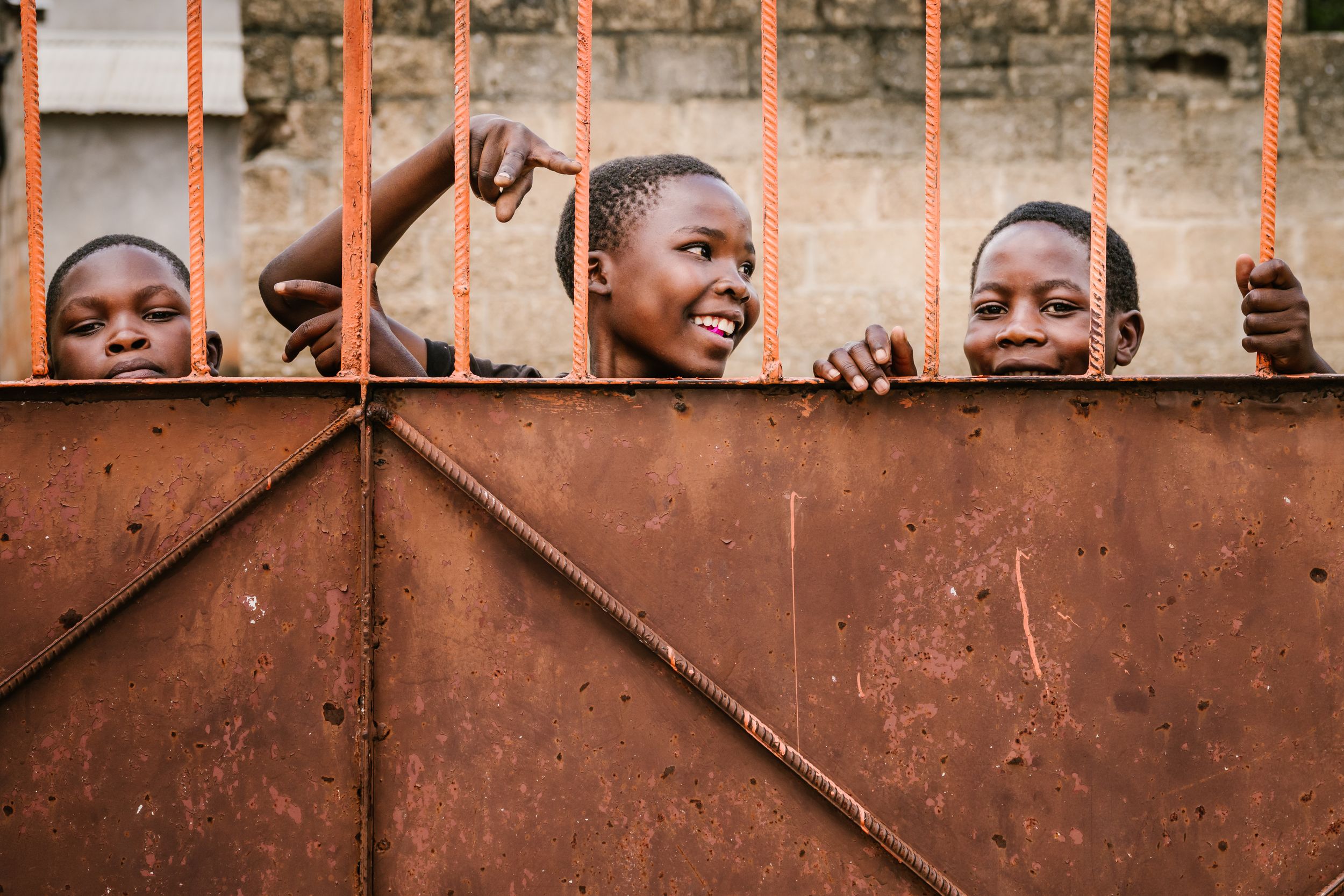 Three smiling African children peering through a rusty metal gate with orange bars.