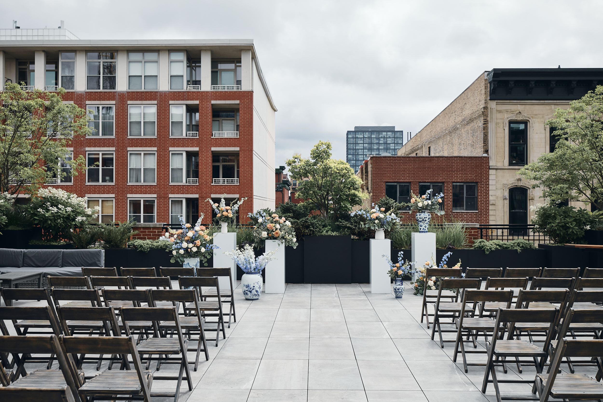 Elegant rooftop wedding ceremony setup with floral arrangements, dark wooden chairs, and urban brick building backdrop.