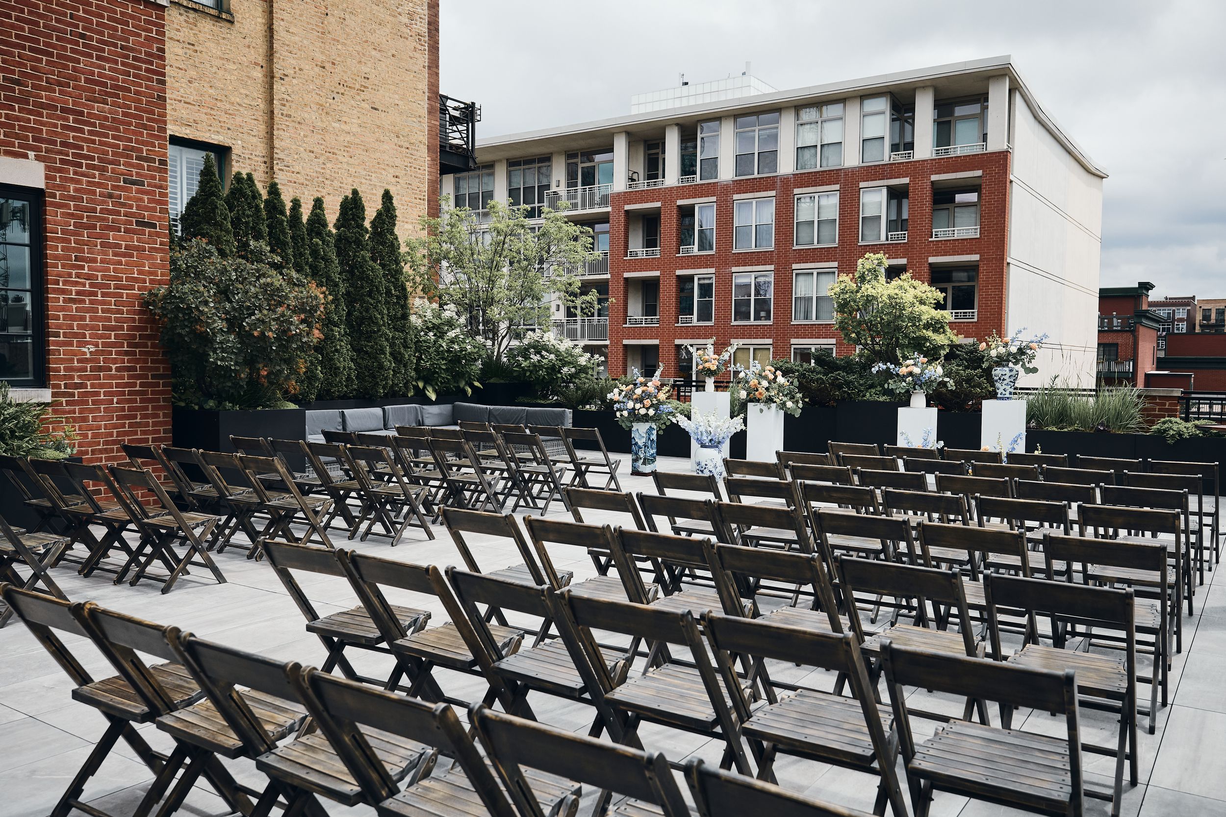 Rows of dark wooden folding chairs arranged outdoors for a wedding ceremony with brick buildings in the background.