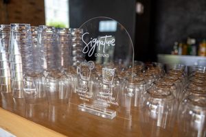 Clear glass jars and an acrylic signature drinks sign displayed on a wooden bar counter at a wedding reception.