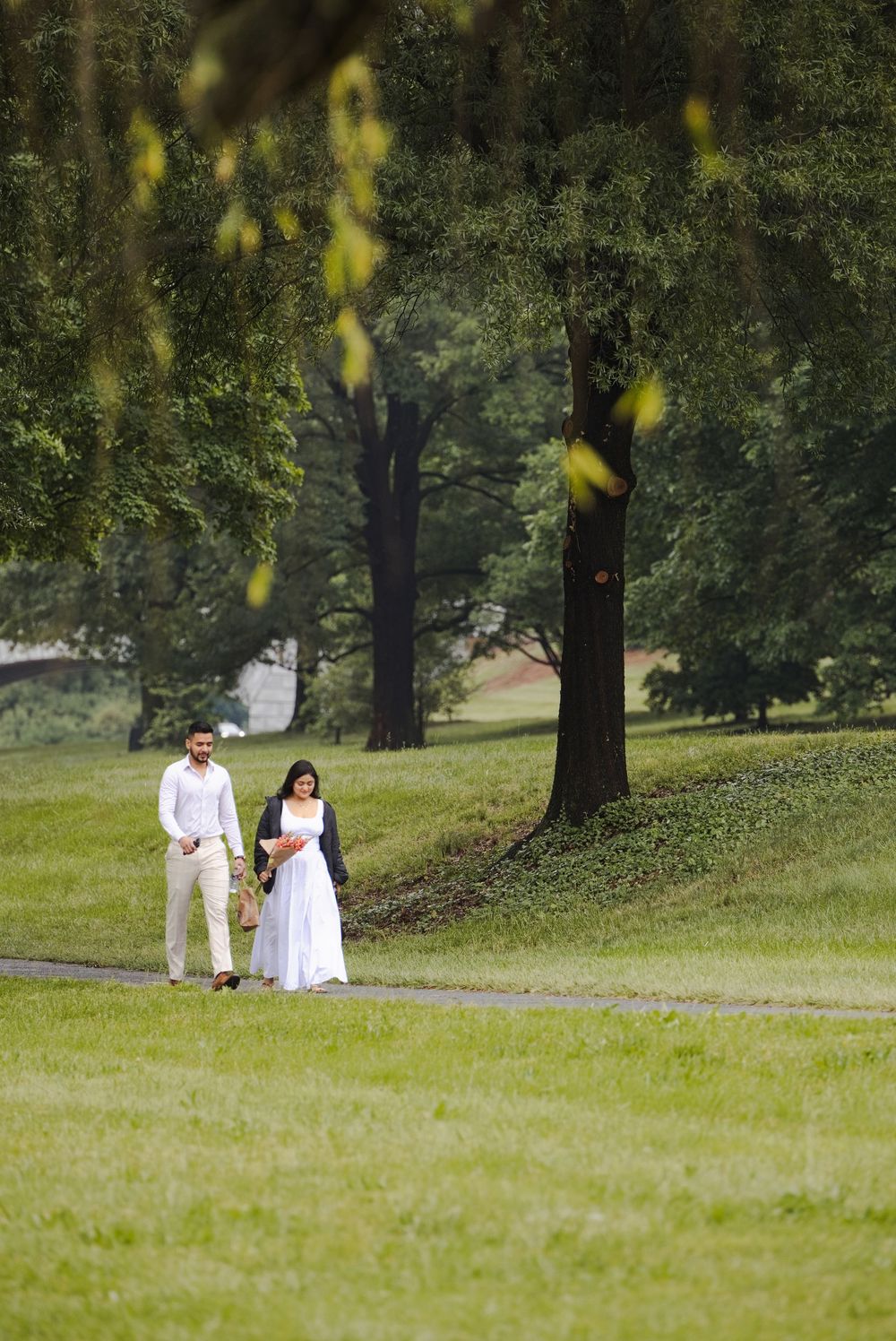 A couple in wedding attire walks hand in hand through a lush green park surrounded by tall trees.