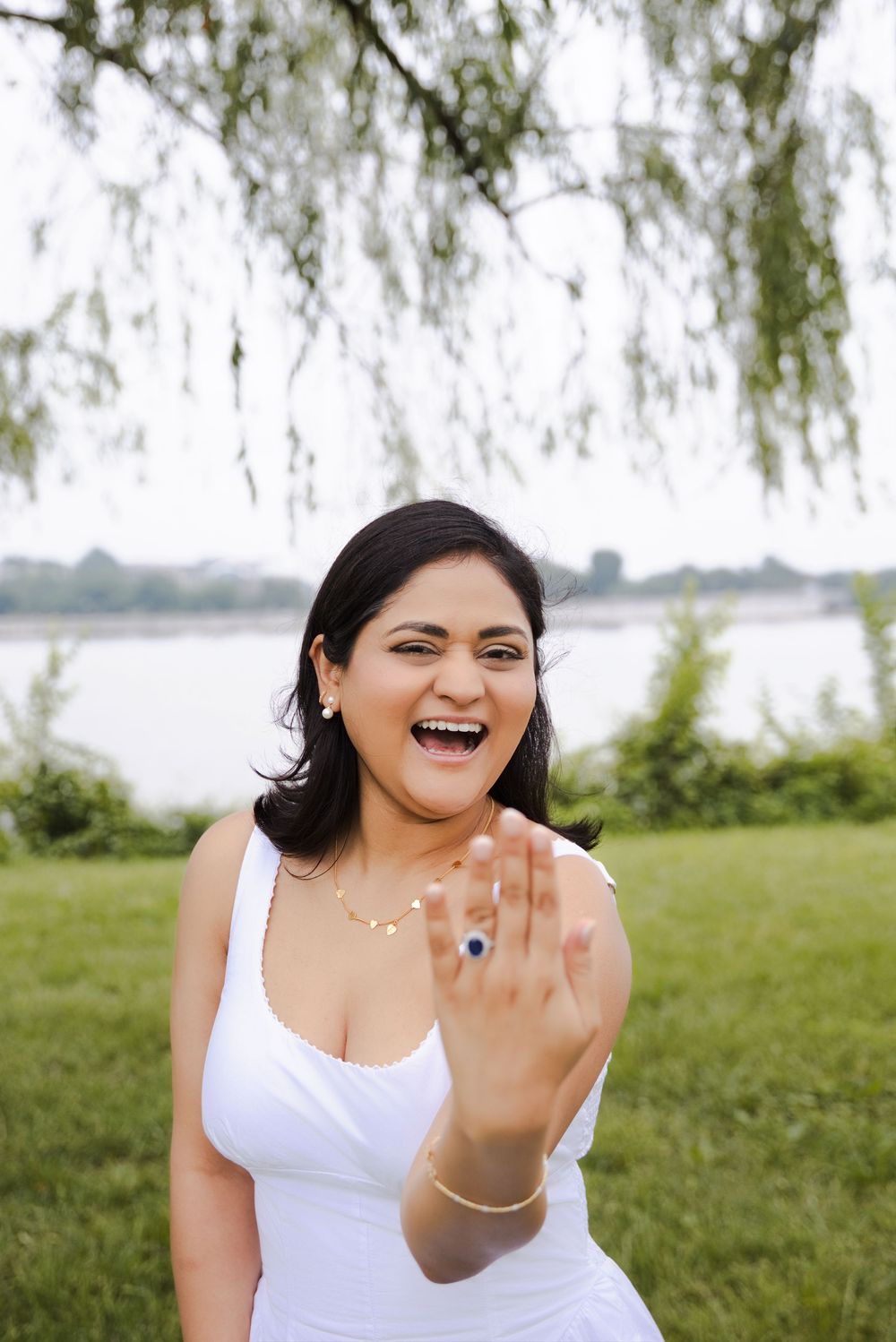 A joyful woman in a white dress shows off her engagement ring outdoors near a river, laughing with excitement.