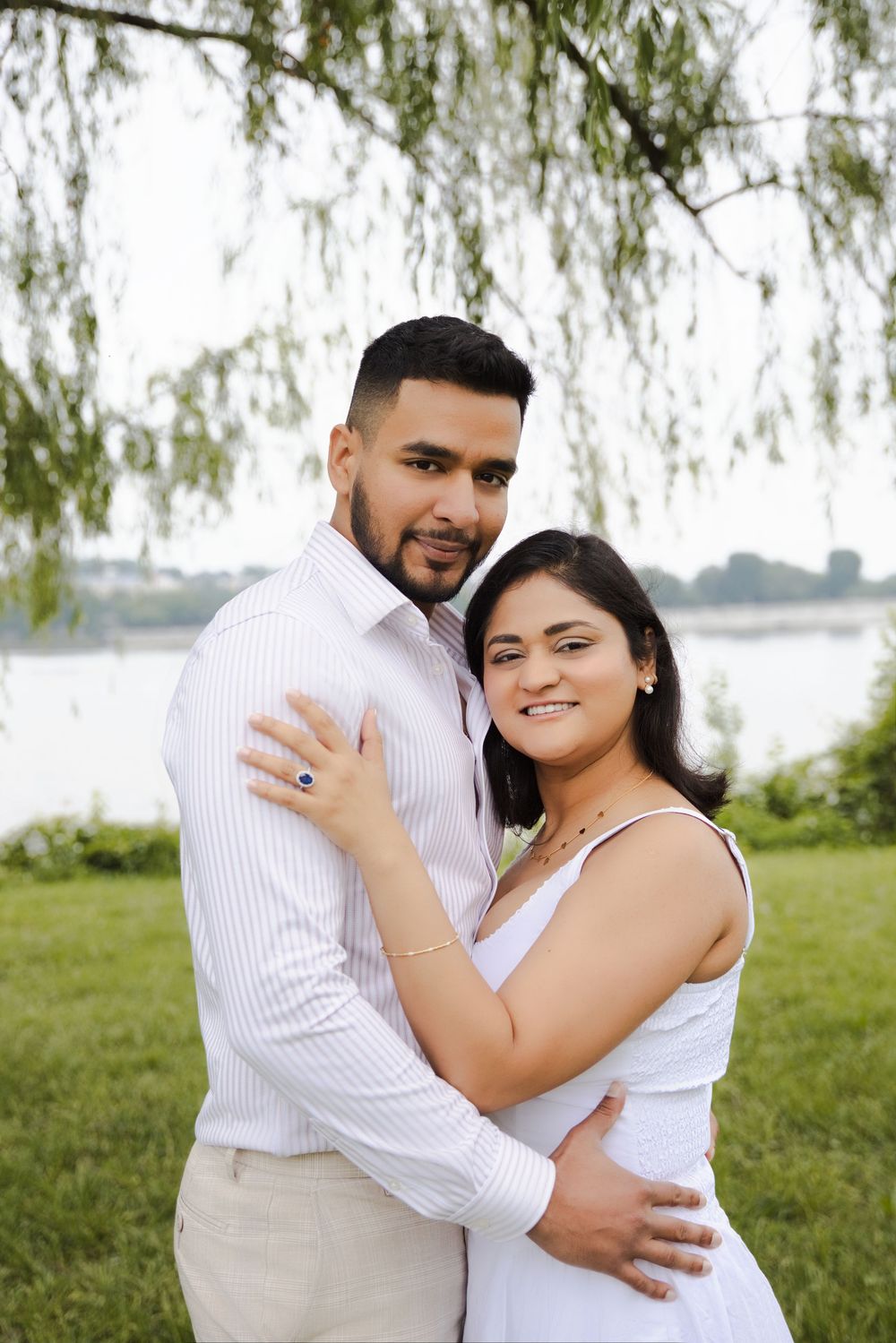 Couple embracing outdoors near a waterfront, woman in white dress with blue ring, man in striped shirt, green trees background.