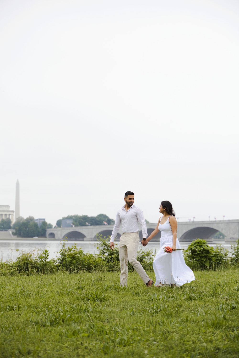 Couple in white attire holding hands walking by the Potomac River with the Washington Monument in the background.