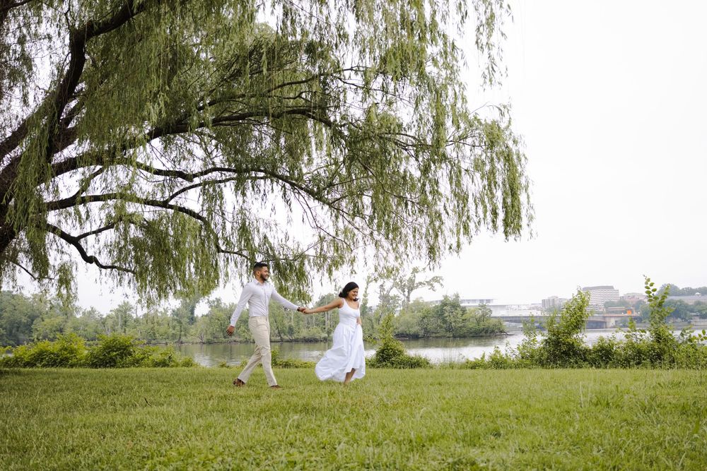 A couple in white attire holds hands and dances joyfully under a weeping willow tree by a serene riverside.