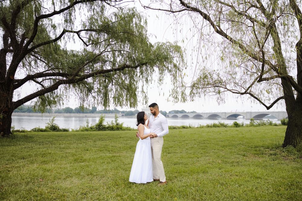 Couple in white attire embracing under willow trees by a serene river with a bridge in the background.