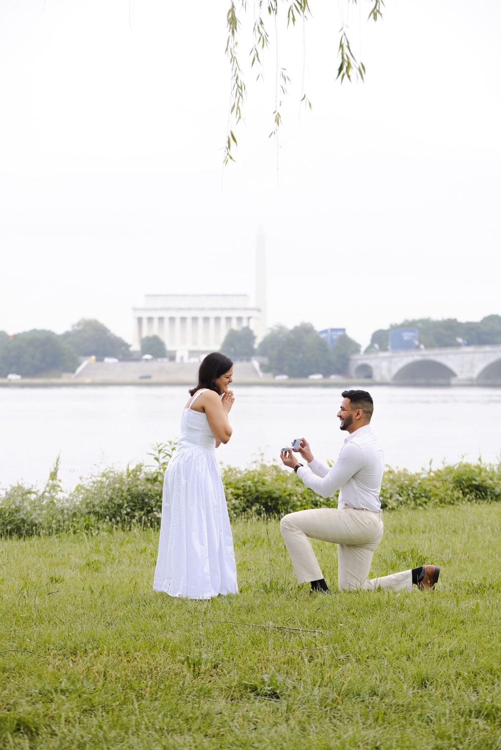Man proposes on one knee to woman in white dress by the Potomac River with Lincoln Memorial backdrop in Washington DC.