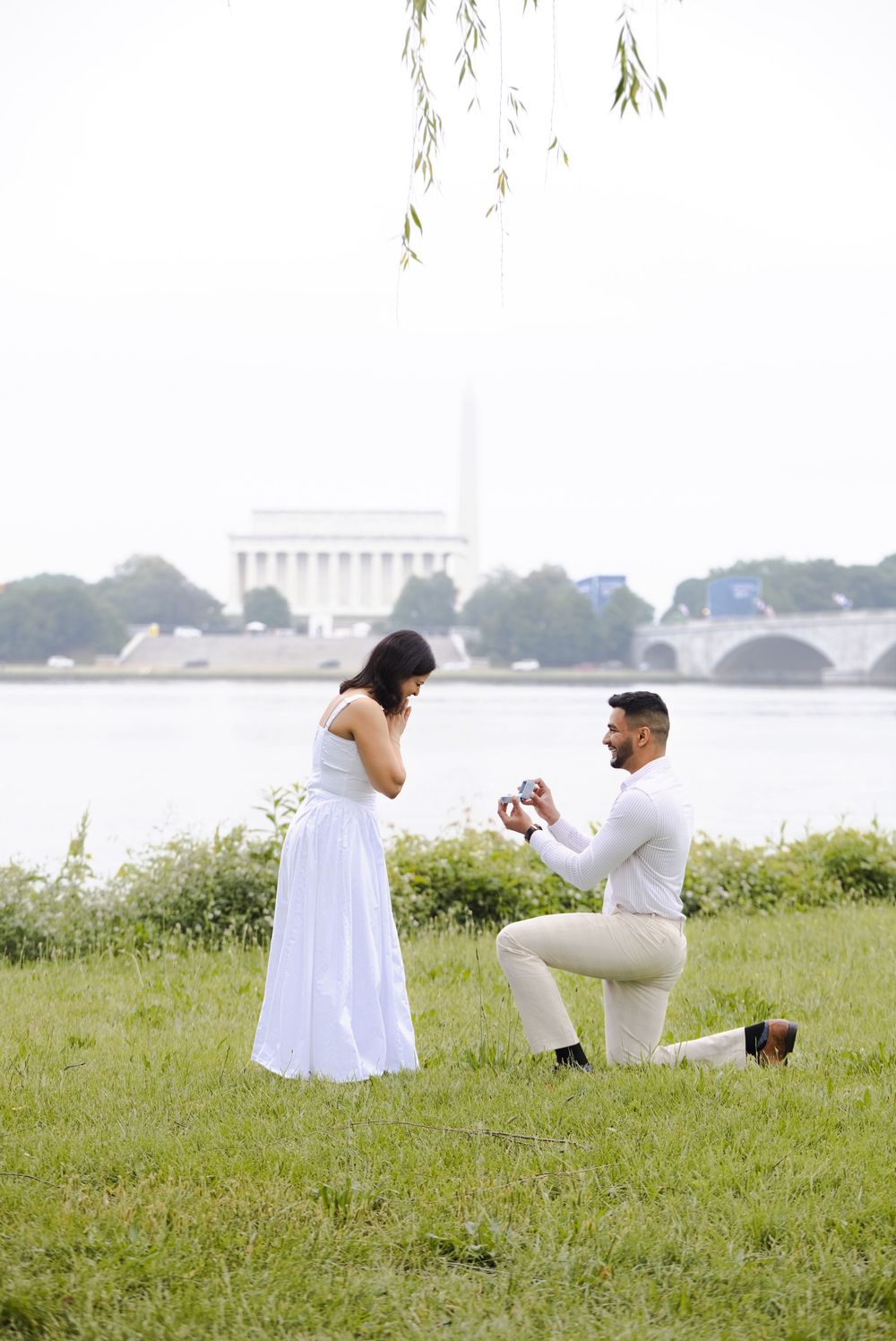 Man proposing on one knee to woman in white dress by the Potomac River with Lincoln Memorial in background.