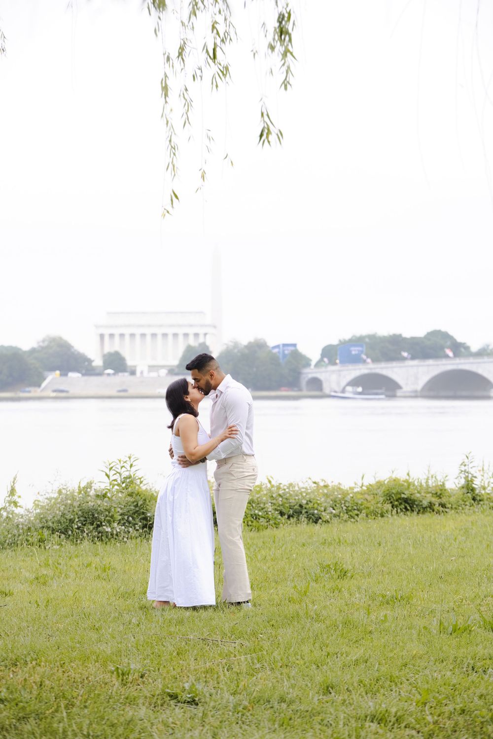 Couple sharing a romantic kiss by the Potomac River with the Lincoln Memorial and Arlington Memorial Bridge in the background.