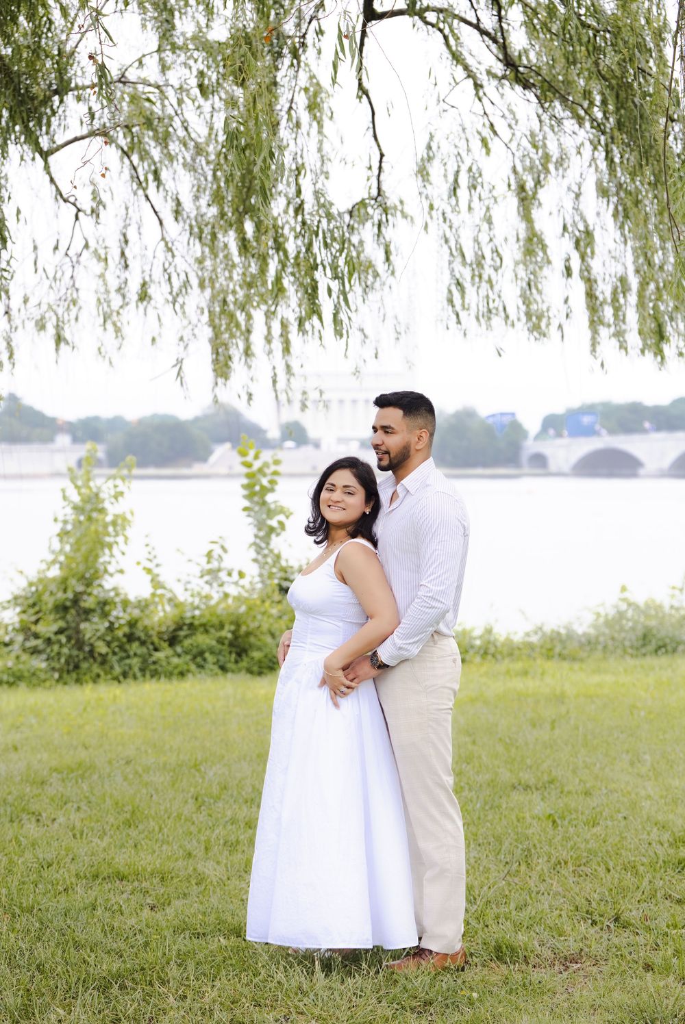 Couple embracing under weeping willow tree by waterfront, woman in white dress, man in white shirt and khaki pants.