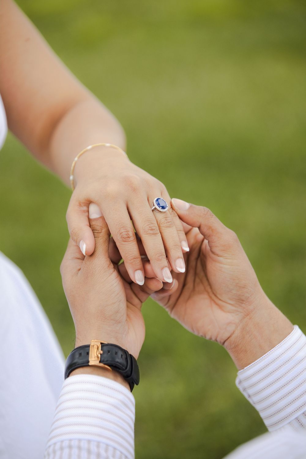 Close-up of couple holding hands with a blue sapphire engagement ring and gold bracelet against green background.