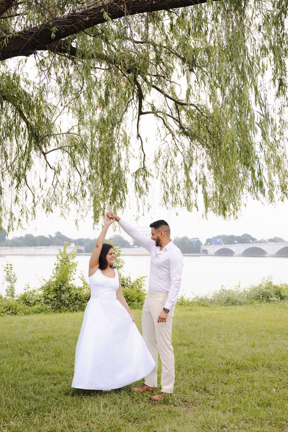 Bride and groom dancing under a weeping willow tree by a river, wearing white wedding attire outdoors.