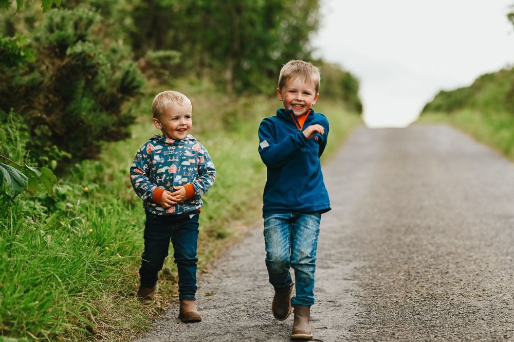 Two young children walk and laugh together on a rural path lined with green foliage.