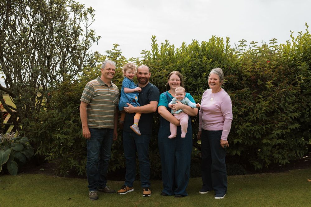 Family of five adults and two babies posing outdoors in front of lush green hedges in a garden setting.