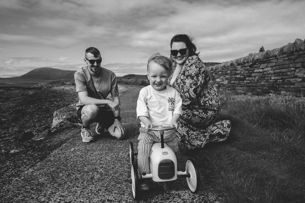 Black and white family photo with parents and toddler on toy car outdoors near stone wall.