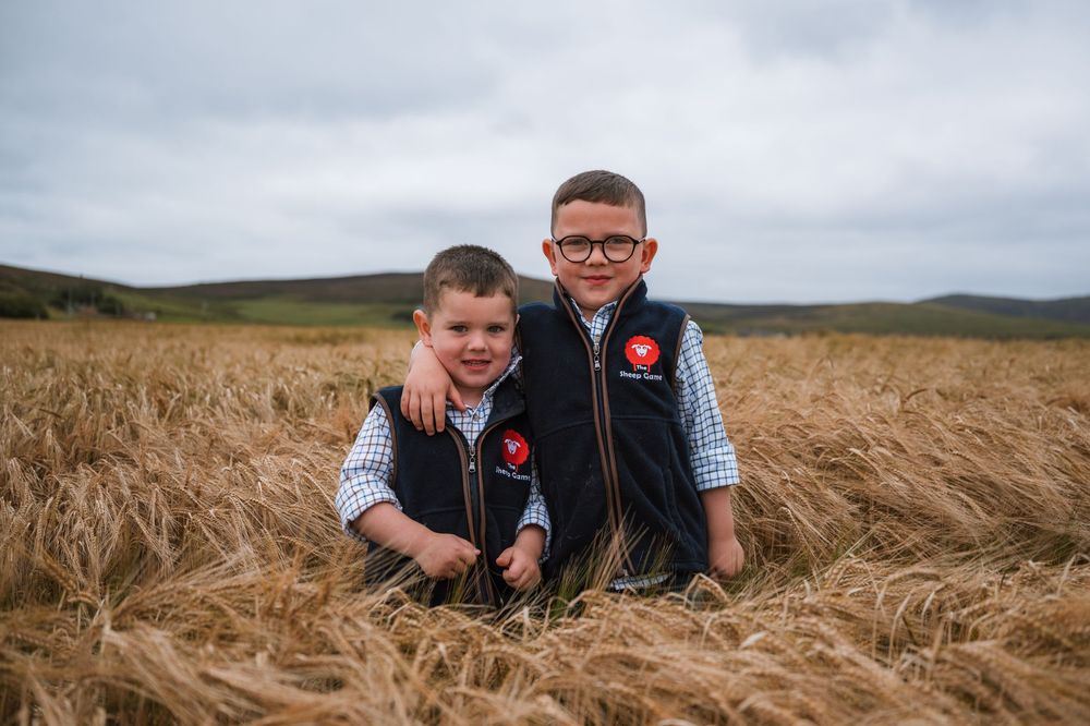 Two young boys in matching navy vests stand smiling in a golden wheat field on a cloudy day.
