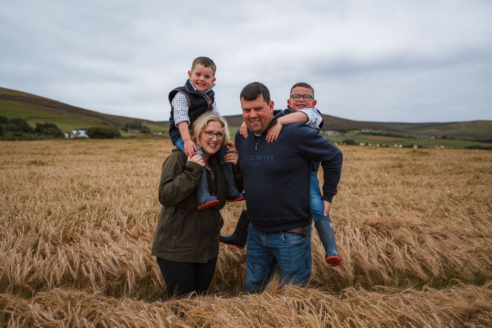 Family of four smiling in a golden wheat field on a cloudy day, two children on parents shoulders.