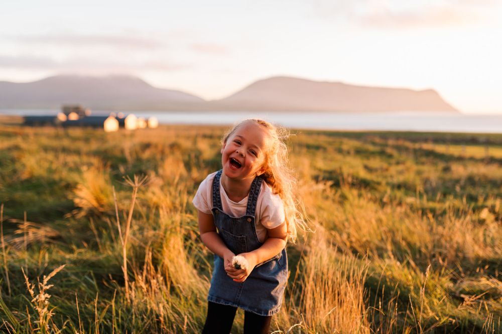 A child laughs joyfully in a sunlit meadow with mountains and farm buildings in the background at golden hour.