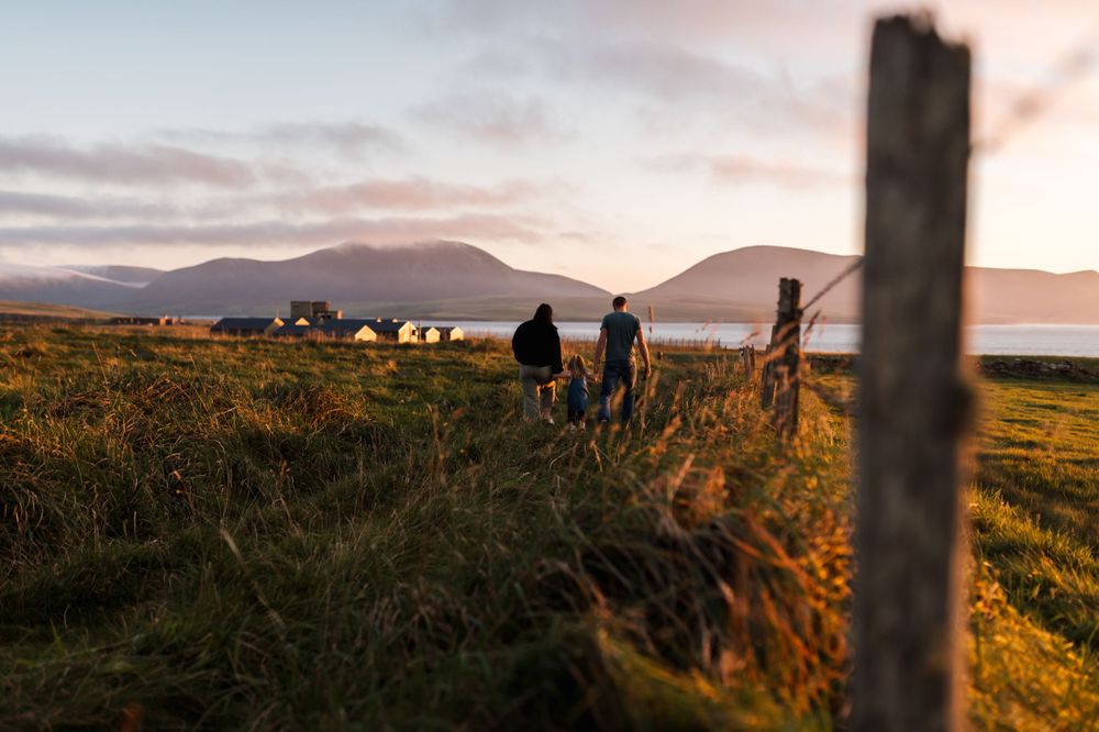 Silhouettes of people walking through a grassy coastal landscape at sunset with mountains in the background.