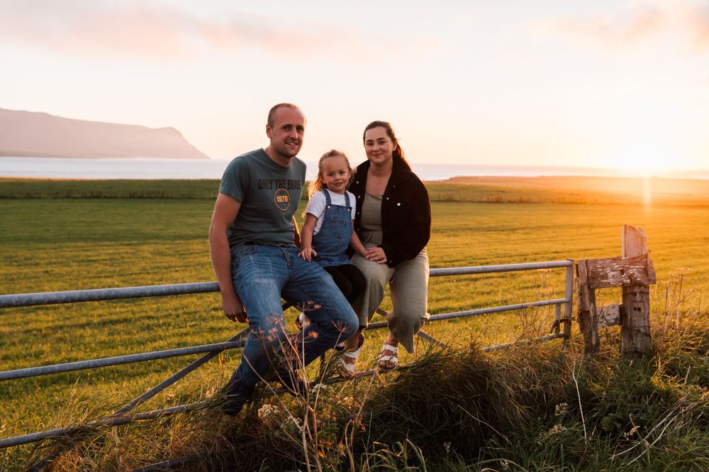 Family posing together on rustic wooden fence at sunset with golden countryside landscape in background.