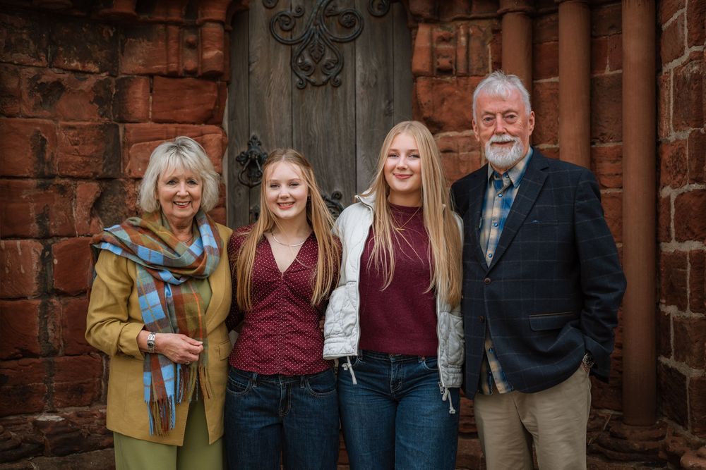 Family of four posing outdoors near a rustic wooden door and brick wall, two teenage girls with older couple.