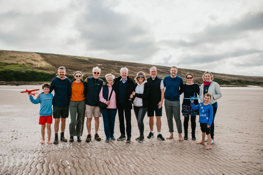 Group of people standing together on a sandy beach with hills in the background under cloudy skies.