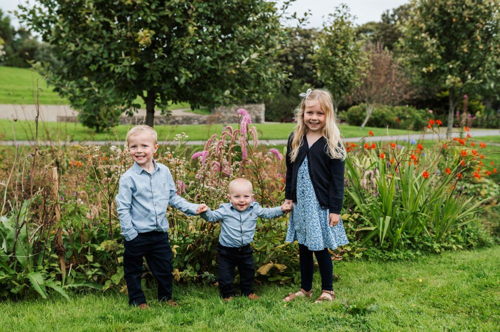 Three young children stand together on green grass with colorful garden flowers and trees in the background.