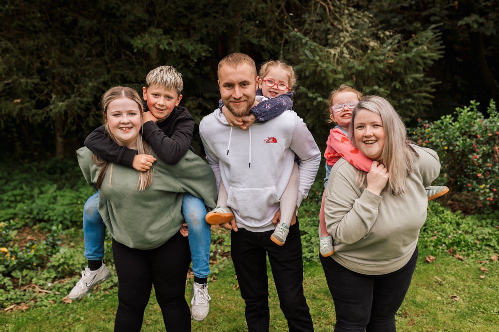 A family group wearing casual clothing poses together outdoors on grass with trees in the background.