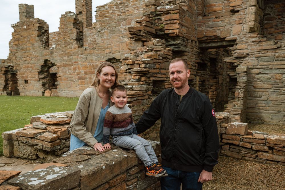 Family of three posing at ancient stone ruins, parents smiling with young child seated on historic wall.