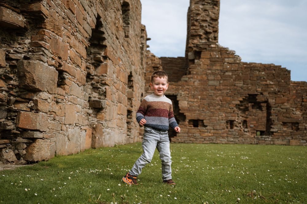Young boy running on grass beside ancient stone castle ruins under a cloudy sky.
