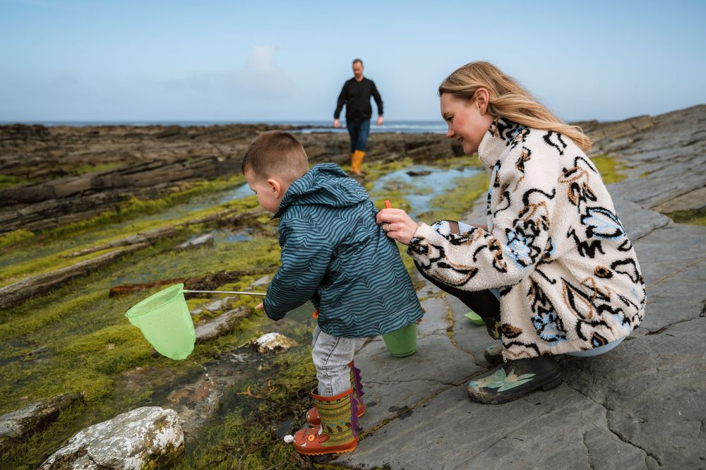 A mother helps her toddler explore rocky tide pools while a man stands in the background on a coastal shoreline.