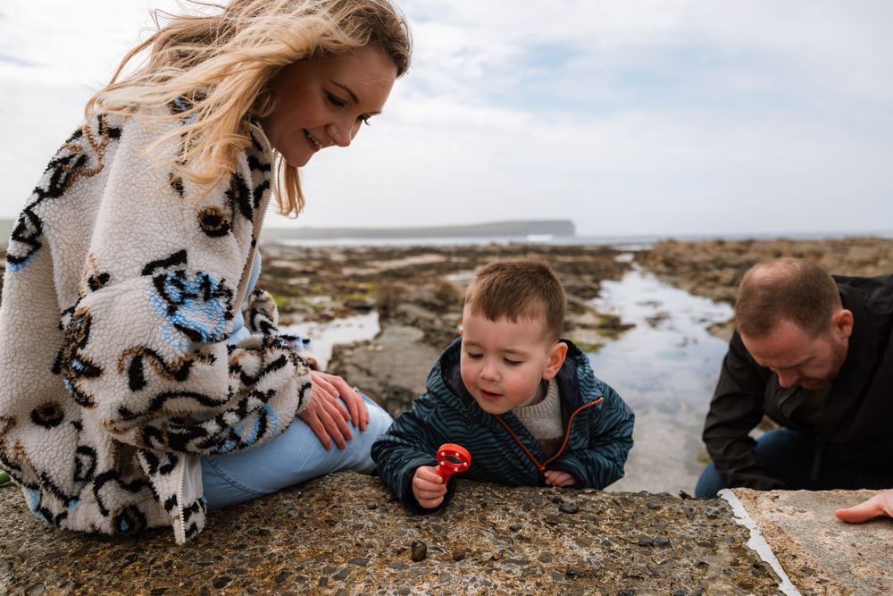 Family with toddler exploring rocky shoreline, mother in patterned wrap smiling as child examines tide pool finds.