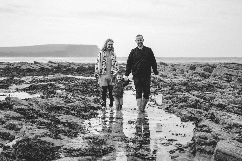 Black and white photo of a couple and child walking on a rocky beach with seaweed and coastal cliffs in the background.