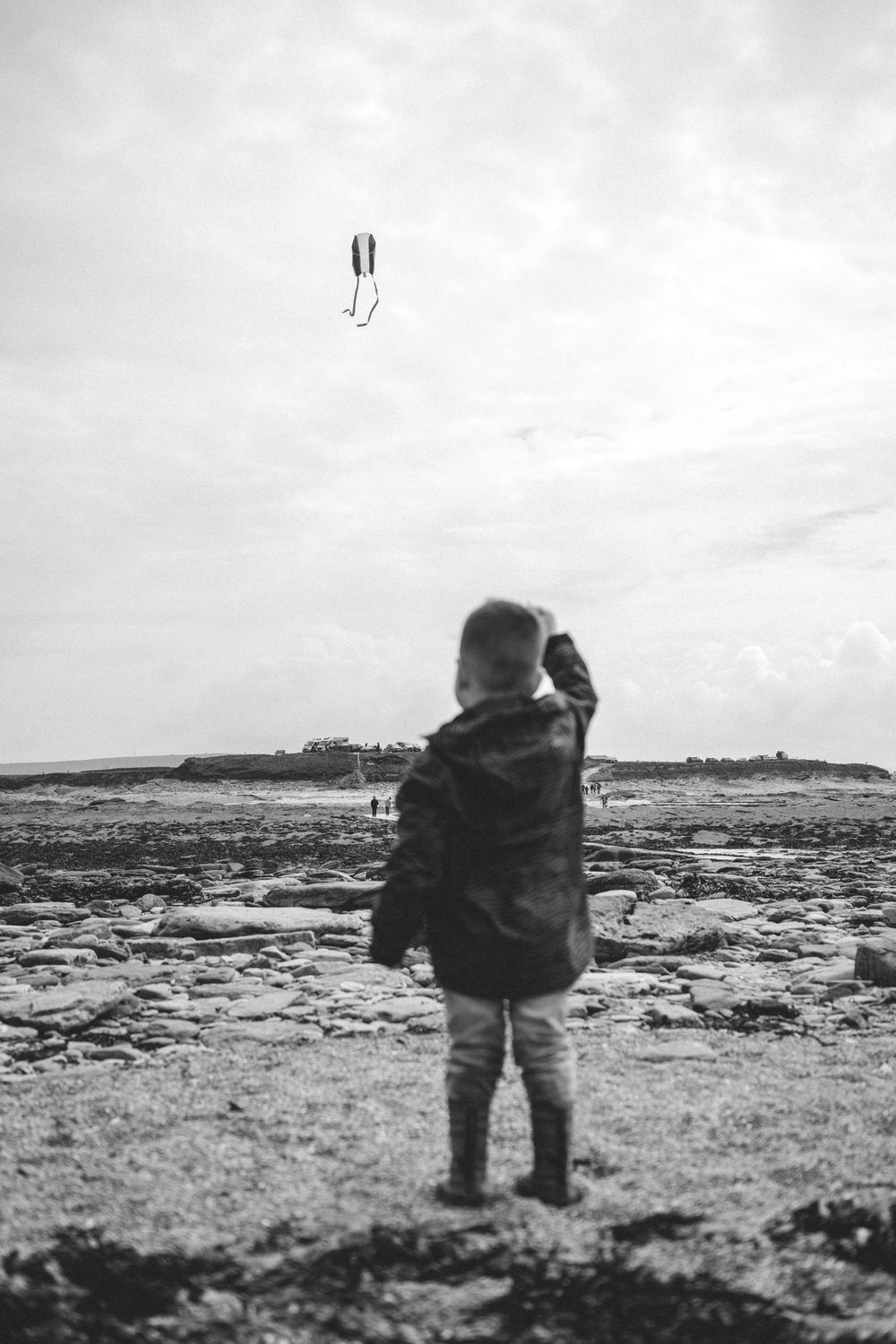Black and white photo of a child flying a kite on a rocky beach, viewed from behind.