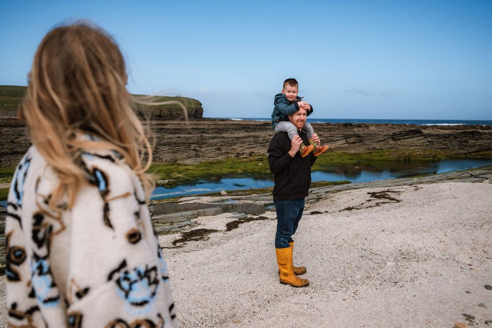 Family enjoying a coastal beach day, father lifting child with daughter watching, rocky shoreline backdrop.