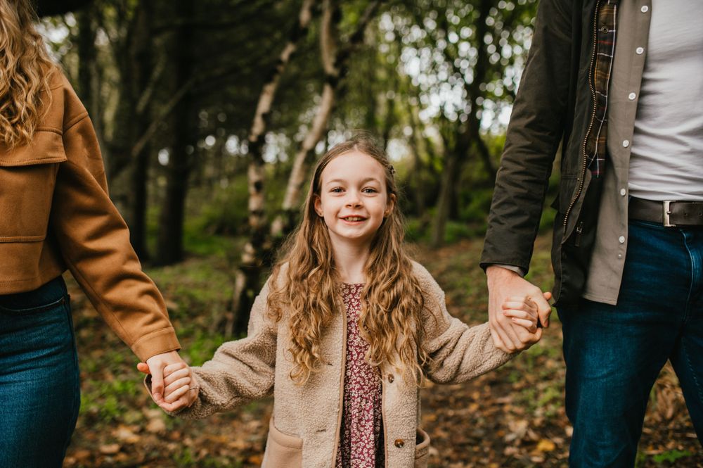 A child in a beige coat walks through autumn leaves while holding hands with family members in a forest.
