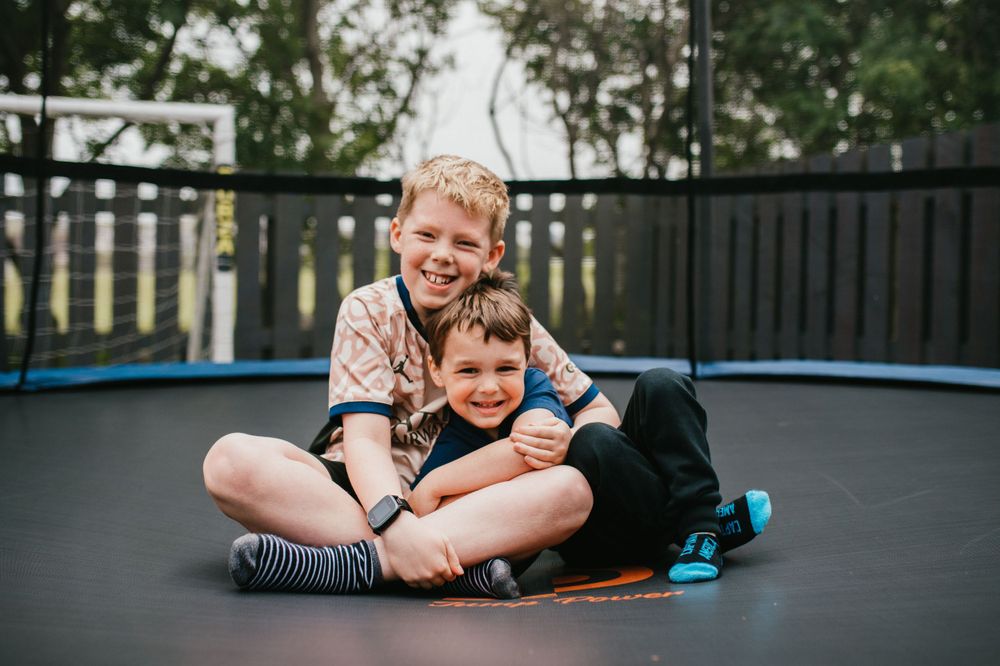 Two young children laughing and hugging while sitting on a black trampoline in a backyard setting.