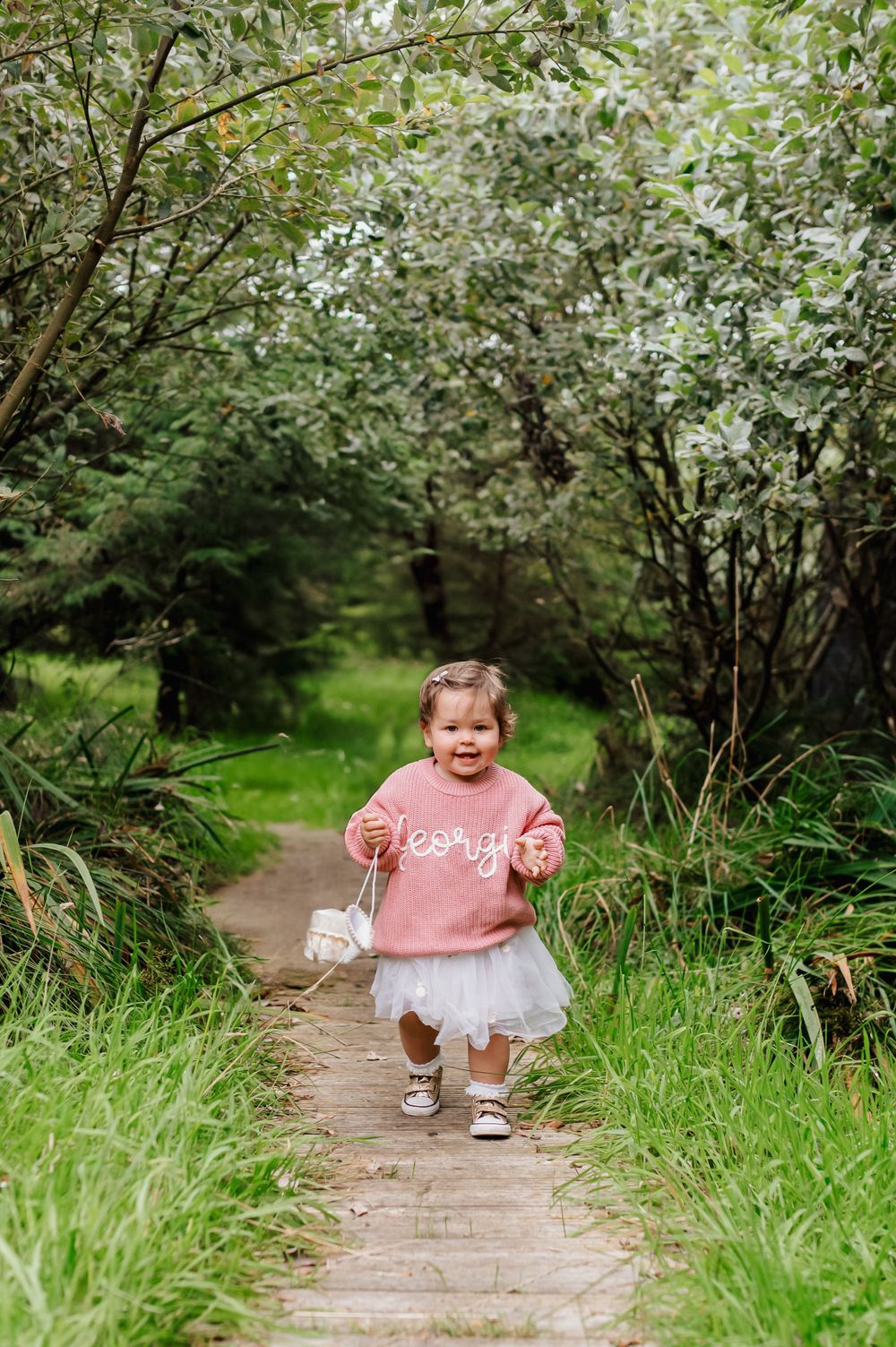 A toddler in a pink sweater and tulle skirt walks along a wooded path surrounded by lush greenery.
