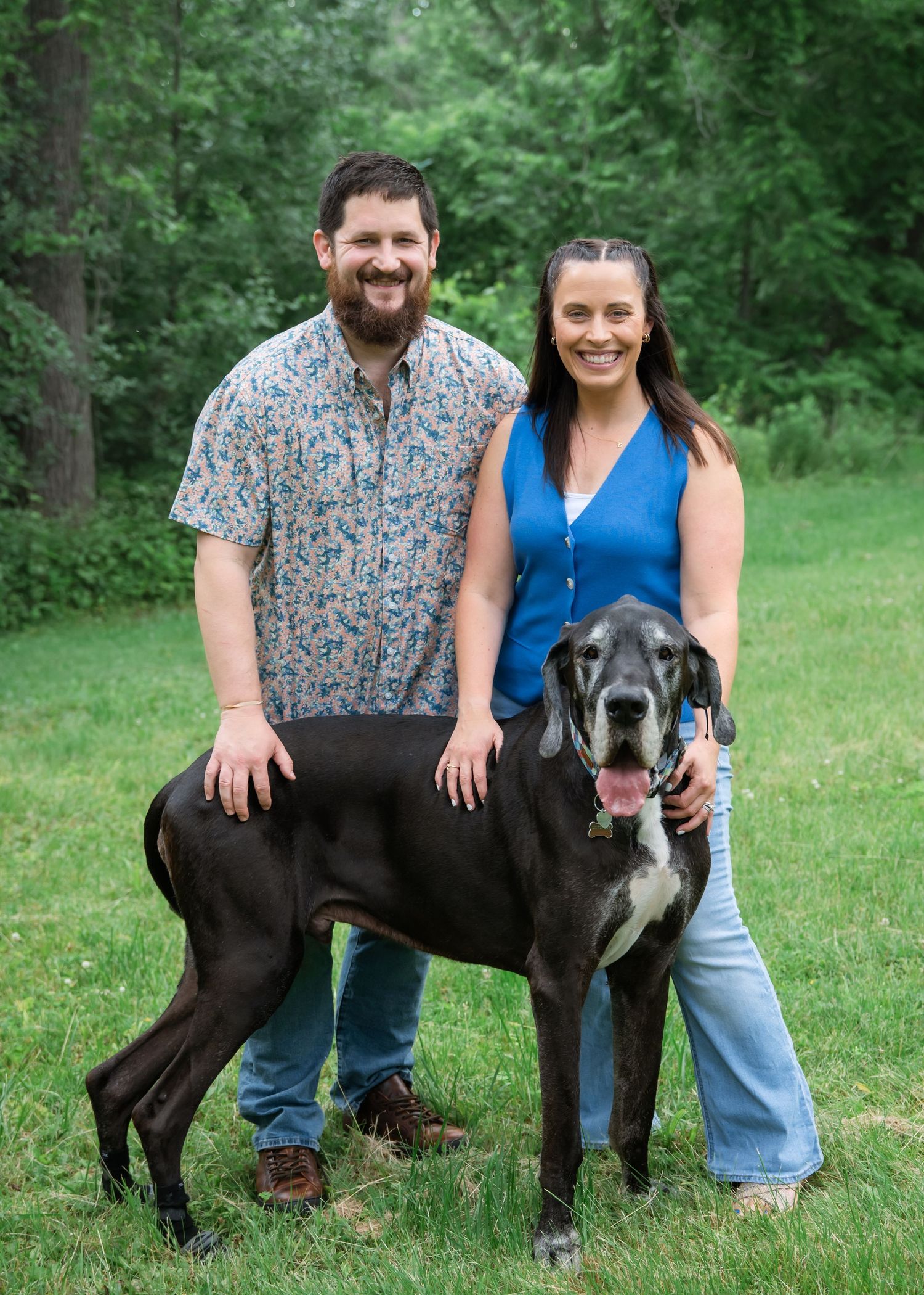 A couple smiling outdoors with a large black Great Dane dog on a green lawn surrounded by trees.